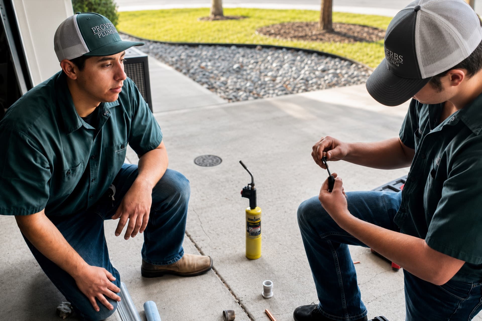 Two Proverbs Plumbing technicians in branded caps and shirts preparing a soldering job on a driveway